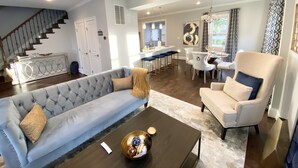 A modern living room with a grey sofa, a coffee table, and a dining area with a white chair and bar stools.