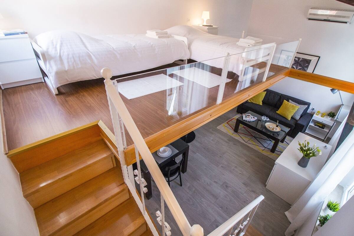 A modern hotel room with a glass balcony, a wooden staircase, a sofa, and a dining area.