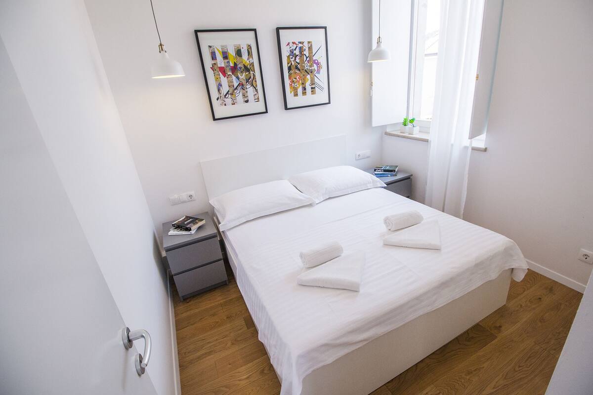 A neatly made bed with white linens, two bedside tables with books, and two framed abstract artworks on the wall.