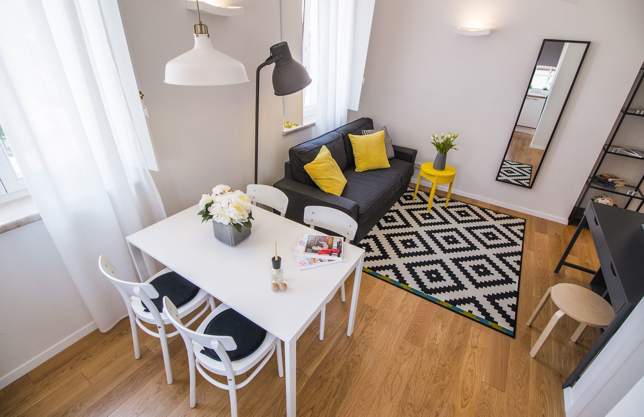 A modern living room with a white dining table, chairs, a grey sofa with yellow cushions, a black floor lamp, a patterned rug, and a small yellow side table with a plant.