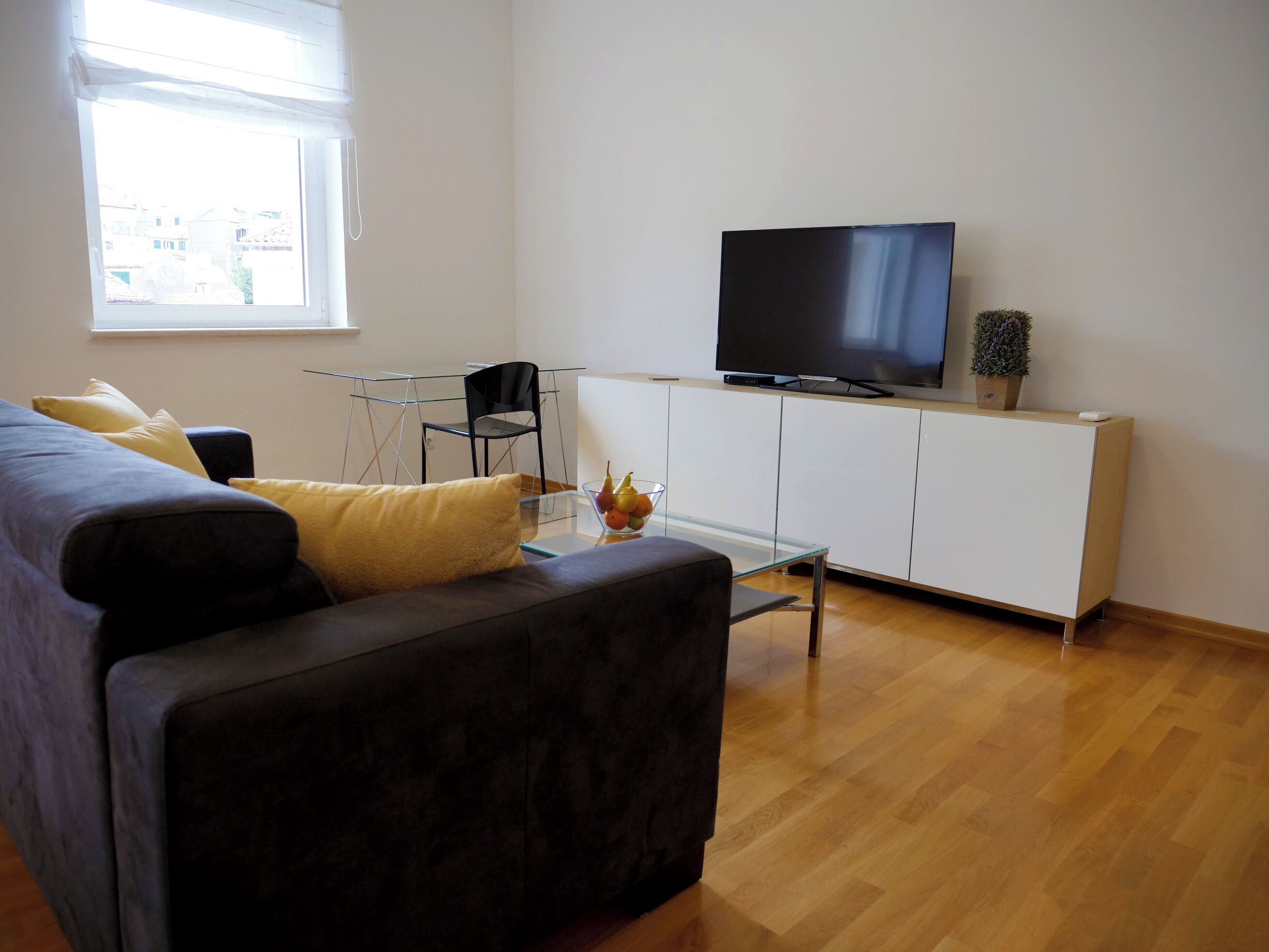 Moderni olohuone, jossa on sohva, lasinen sohvapöytä, valkoisessa kaapissa oleva televisio ja pieni huonekasvi.