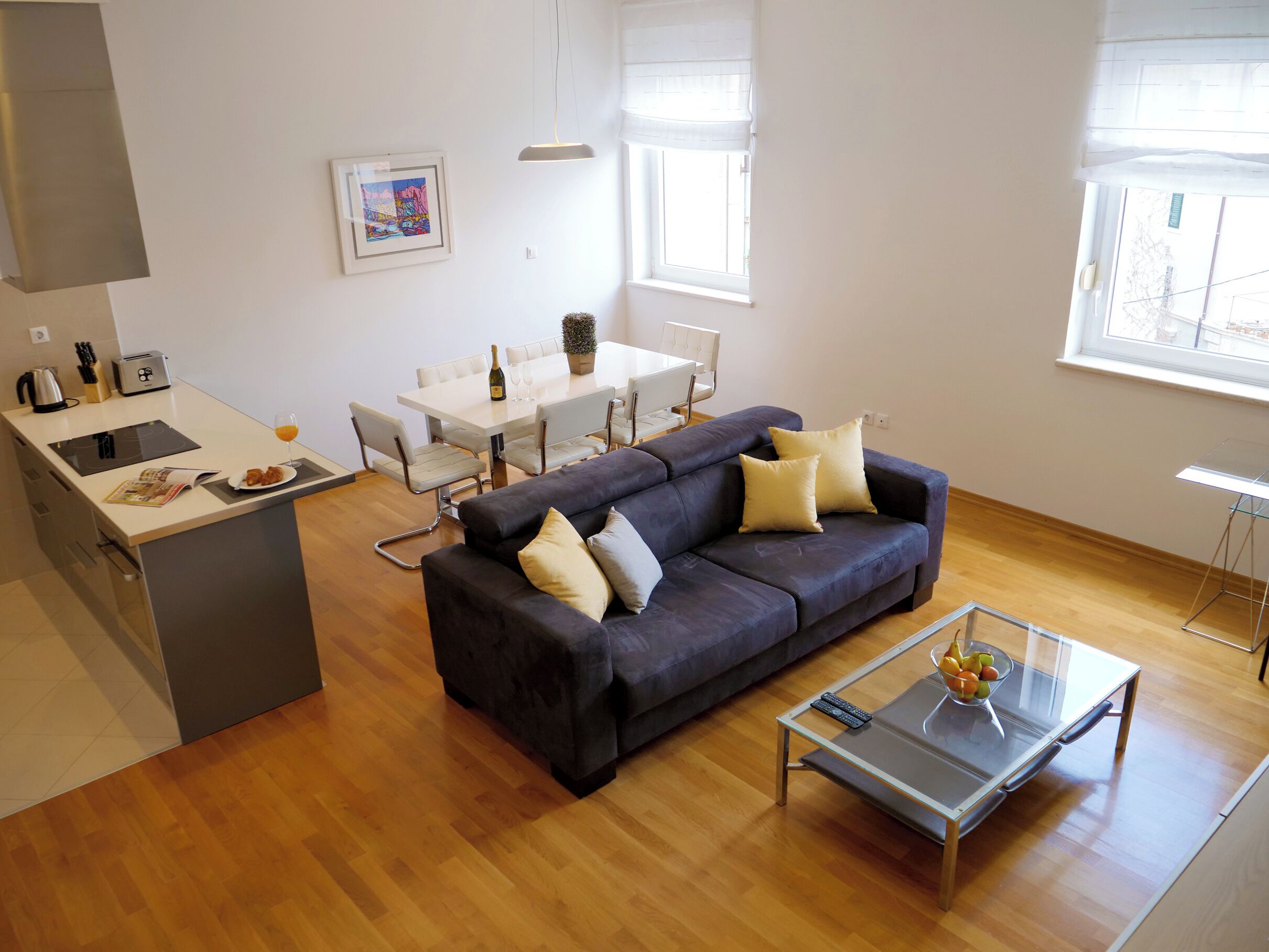 Una sala moderna con un sofá oscuro, una mesa de centro de vidrio y una zona de comedor con una mesa y sillas blancas.