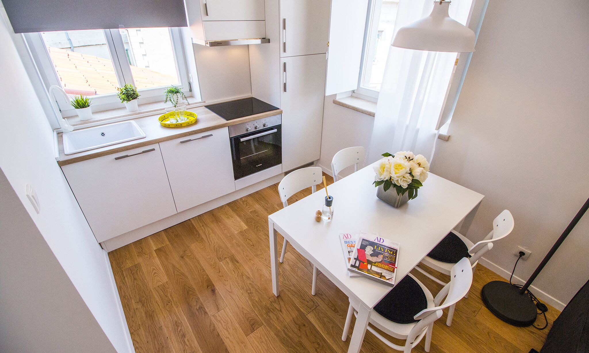 A modern kitchen with white cabinets, a black oven, and a window with plants. There's a dining table with chairs and a magazine.