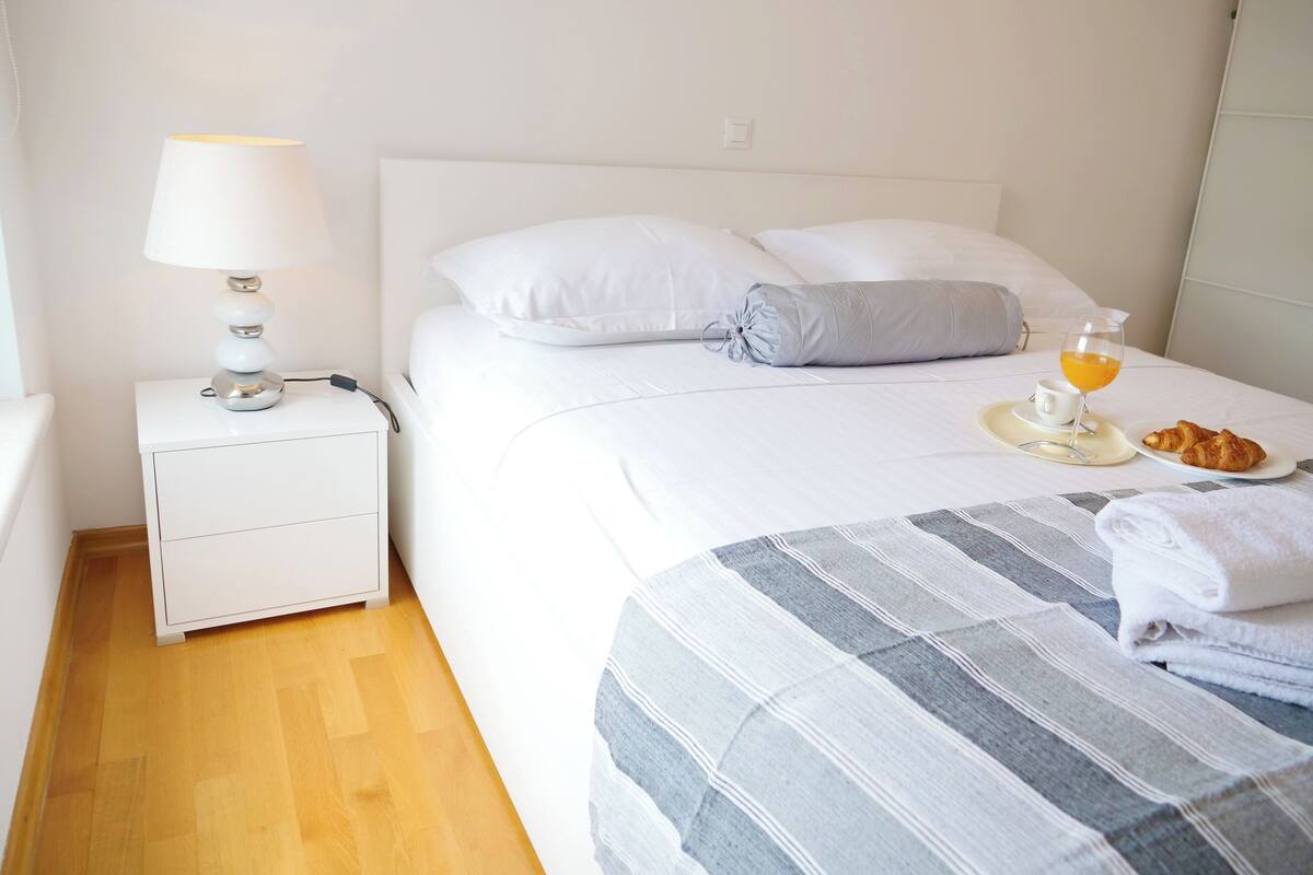 A neatly made bed with pillows, a bedside table with a lamp, a tray with croissants, a glass of orange juice, and a folded towel.