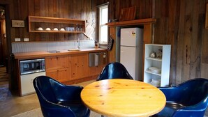 A rustic kitchen with wooden cabinets, a built-in microwave, and a refrigerator. There is a wooden table with two blue chairs and a white shelf with dishes. - Historic Miners Cottage on Howqua River (Howqua)