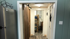 Un pasillo estrecho con una puerta de madera, un cubo de basura y una cama litera al fondo.