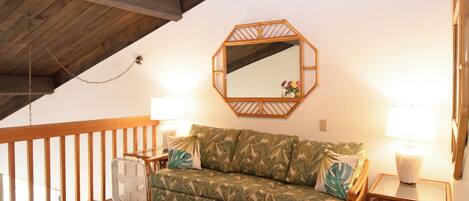 A cozy room with a wooden ceiling, a green patterned sofa, two bedside lamps, and a small table with a lamp.
