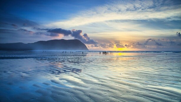 Una playa con un atardecer, aguas tranquilas y una montaña a lo lejos.