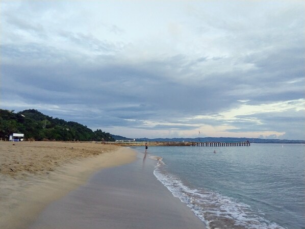 A beach with a sandy shore, a pier extending into the water, and a hilly landscape in the background.