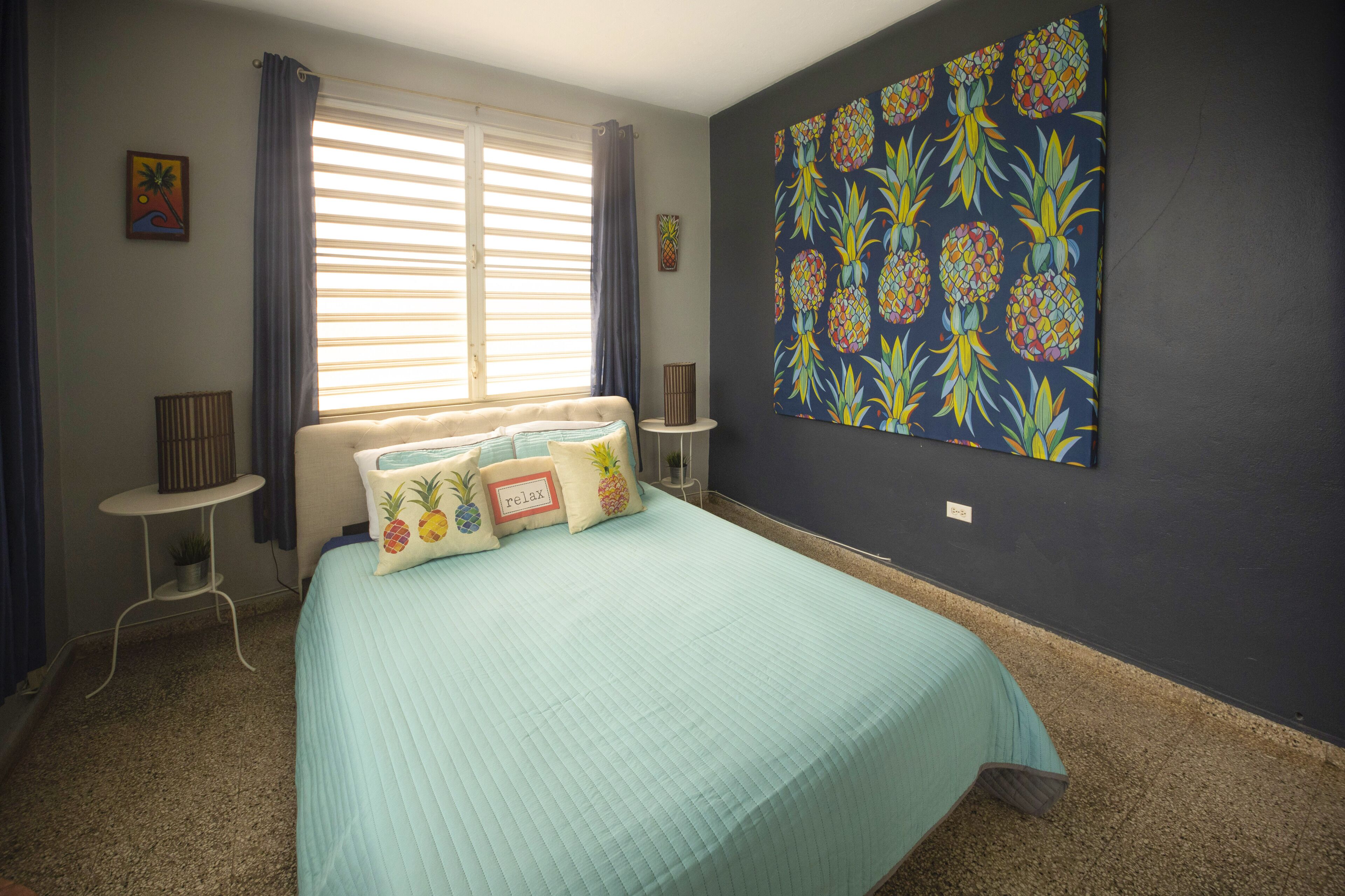 Une chambre à coucher avec un lit, une fenêtre avec des rideaux, un mur orné d’un motif ananas et une petite table avec une lampe.