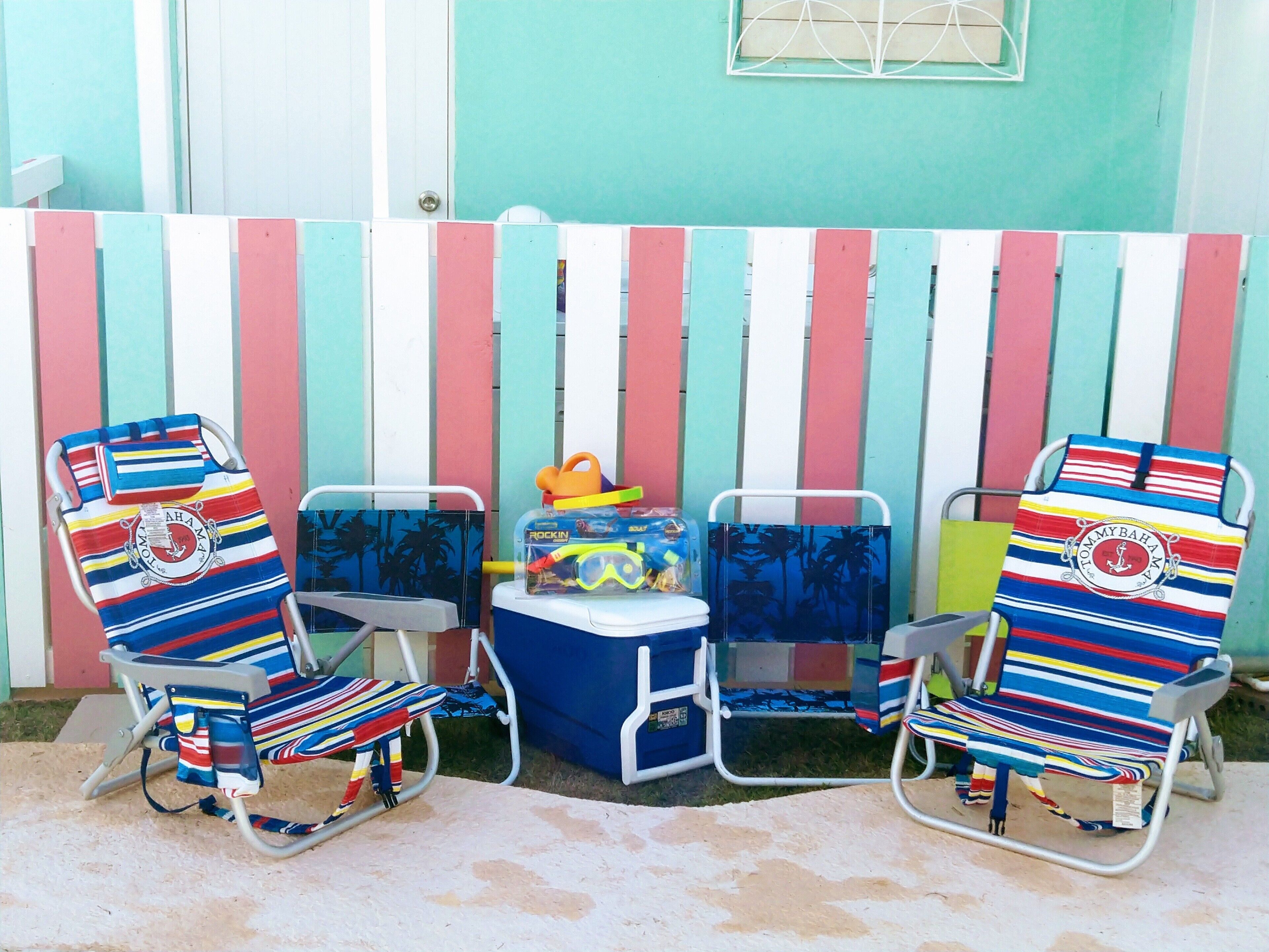 Two beach chairs with striped cushions, a small cooler, and a toy chest against a colorful striped fence.
