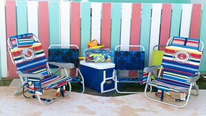 Two beach chairs with striped cushions, a small cooler, and a toy chest against a colorful striped fence.