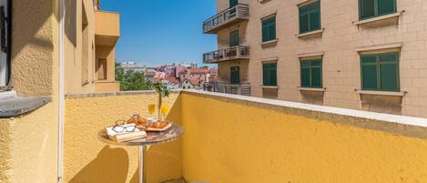 A balcony with a table and two chairs, a small table with a plate of food and drinks, and a view of buildings in the background.