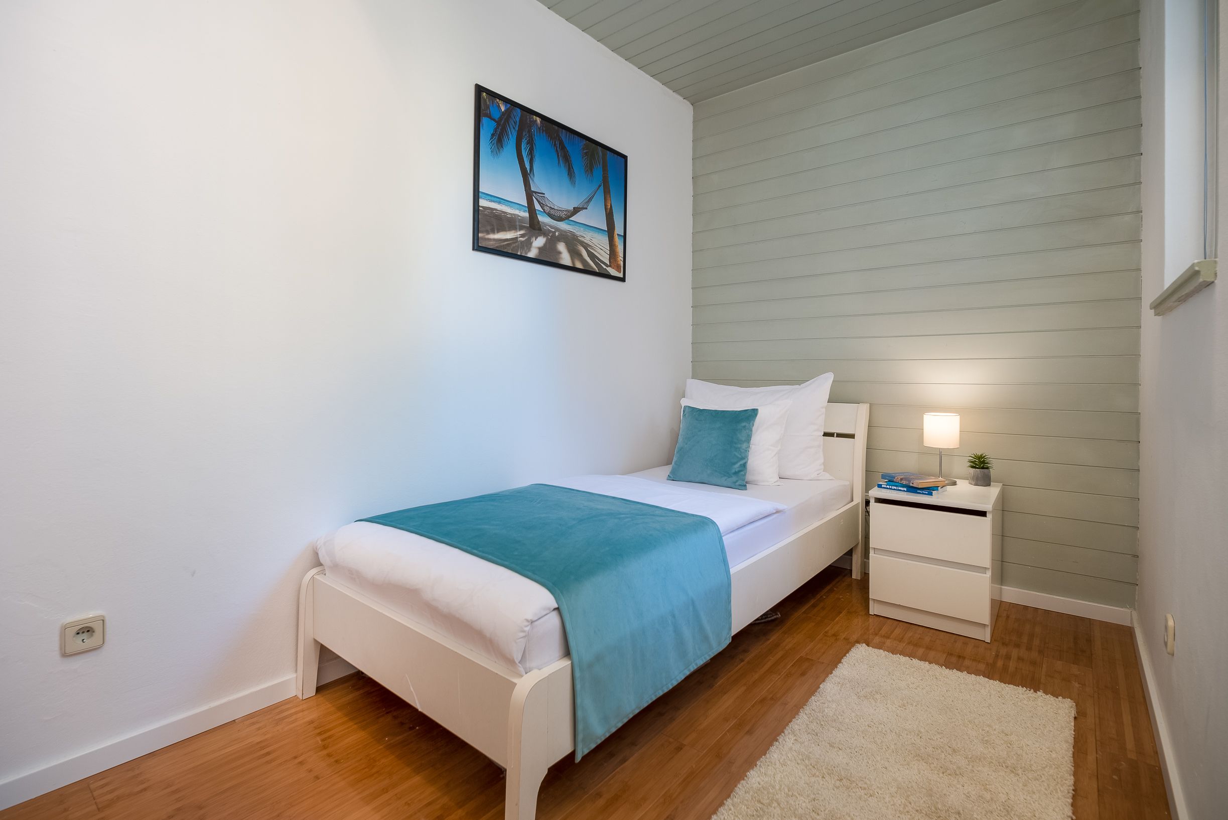 A bedroom with a white bed, teal bedding, a nightstand with a lamp, and a painting of a beach scene.