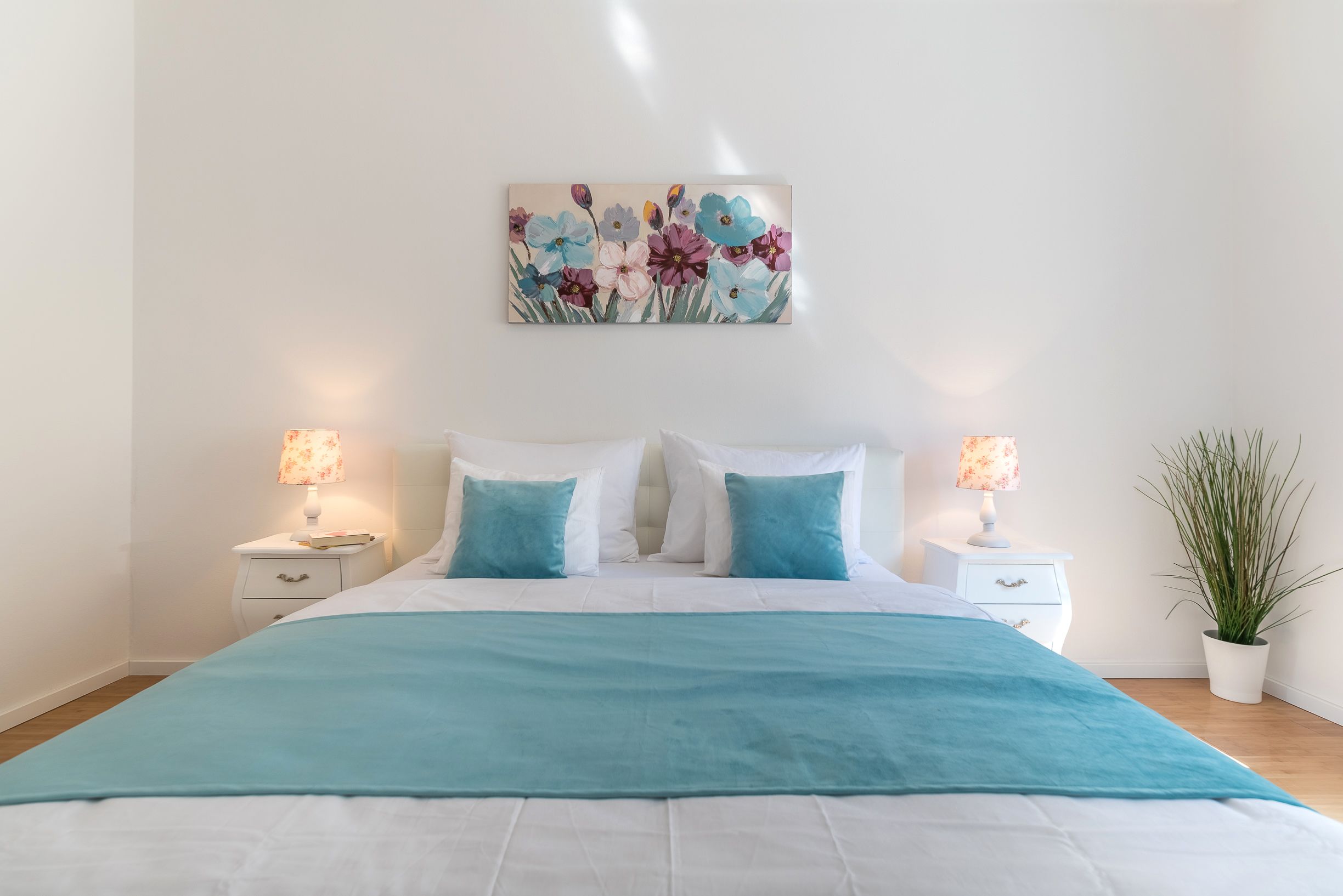 A bedroom with a bed, two bedside tables with lamps, a wall-mounted painting, and a potted plant.