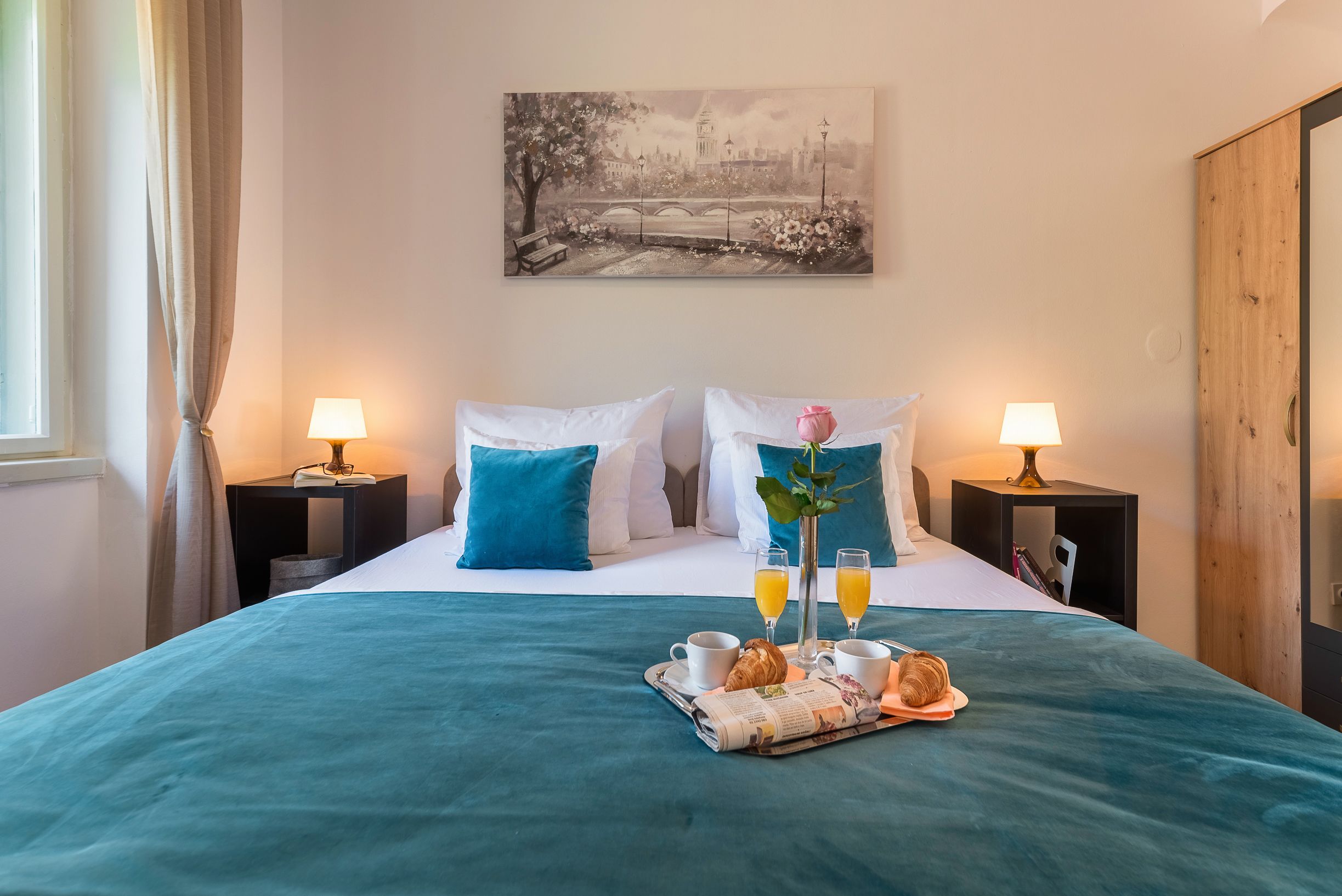 A hotel room with a bed, two bedside tables with lamps, a painting on the wall, and a breakfast tray with croissants, juice, and a newspaper.