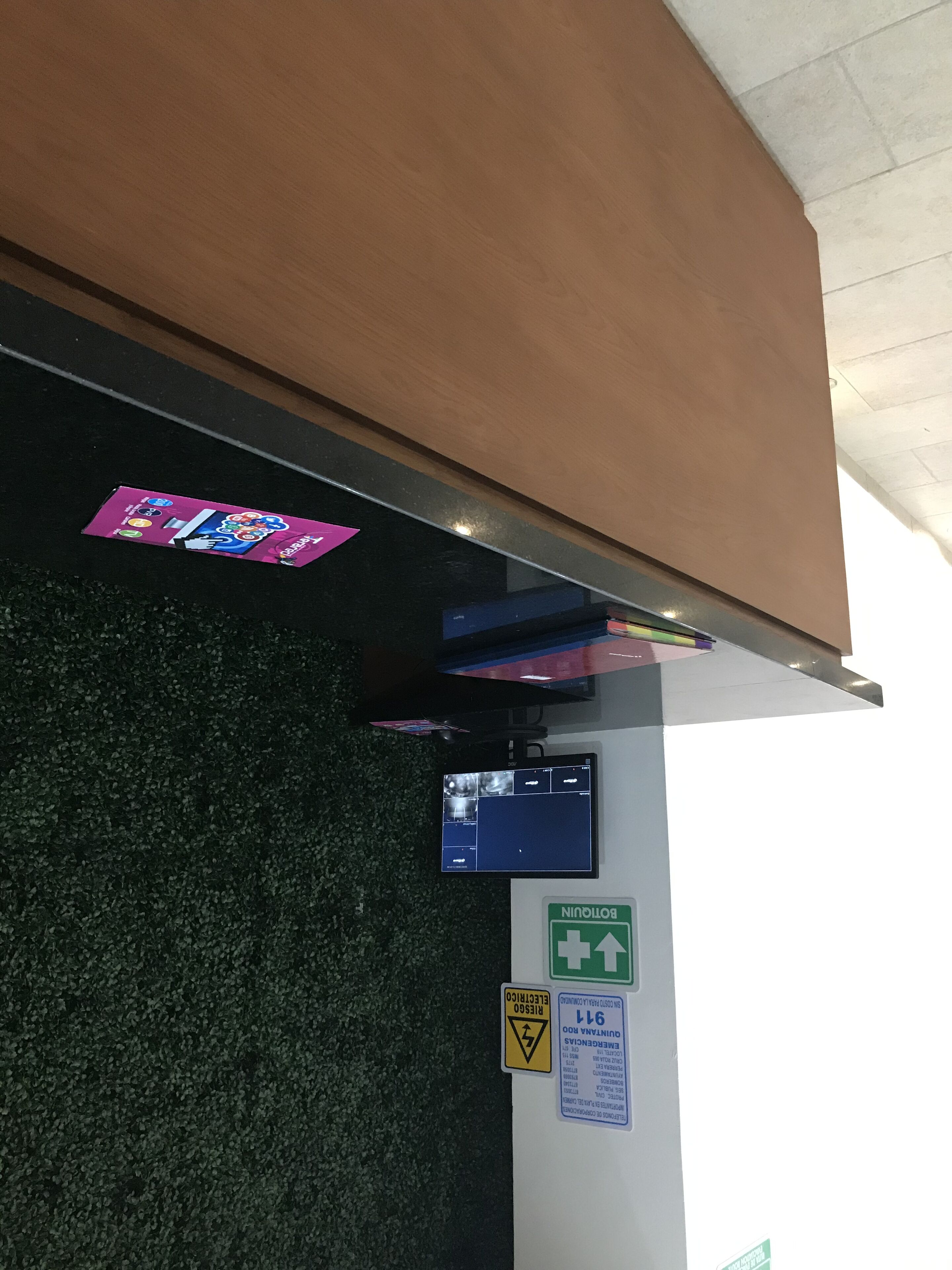 A room with a green emergency exit sign, a warning sign for electrical hazards, and a digital screen displaying room information.