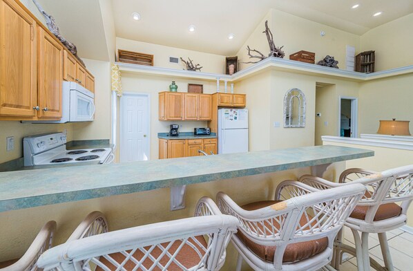 Una cocina con gabinetes de madera, una estufa blanca, un refrigerador y una zona de comedor con sillas de mimbre.