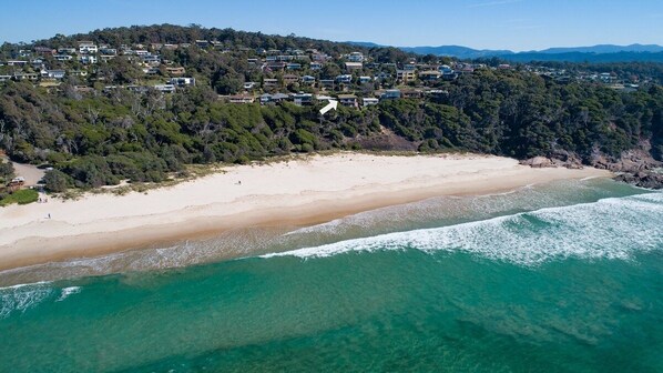 An aerial view of a coastal area with a sandy beach, clear turquoise waters, and a densely wooded hillside with residential buildings.