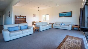 A spacious living room with two light-colored sofas, a wooden bookshelf, a dining table with chairs, and a large window providing natural light. - Unit 3 Beachside units.  Pambula Beach (Pambula Beach)