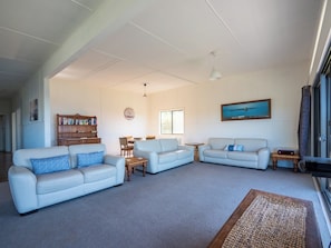 A spacious living room with two light-colored sofas, a wooden bookshelf, a dining table with chairs, and a large window providing natural light. - Unit 3 Beachside units.  Pambula Beach (Pambula Beach)