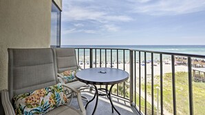 Ein Balkon mit rundem Tisch und zwei Stühlen, der auf einen Strand mit Sonnenschirmen und Menschen blickt.