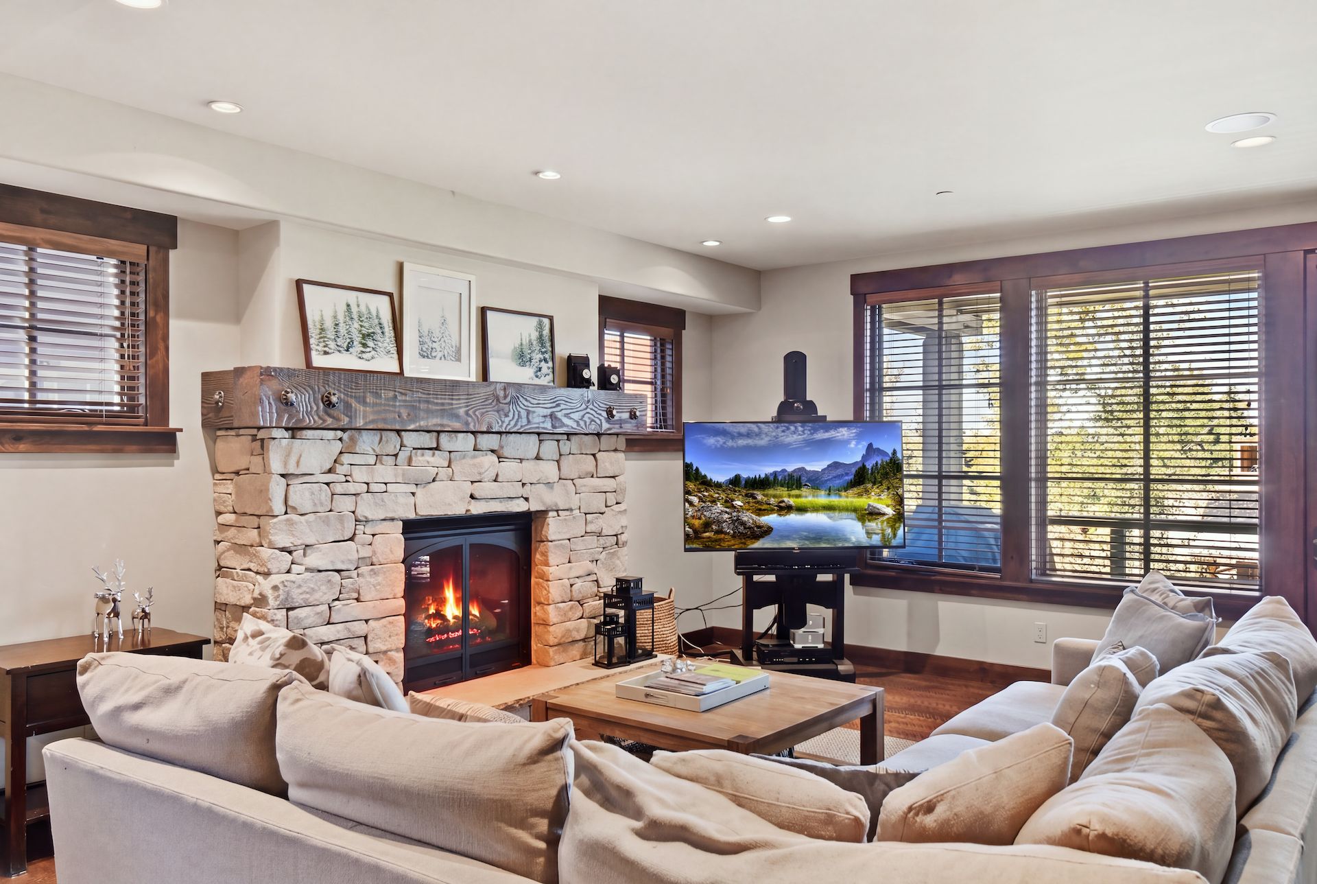 A living room with a stone fireplace, a flat-screen TV, and a large window with blinds.
