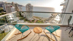 Un balcon avec deux fauteuils à bascule et une petite table, d’où l’on peut admirer un paysage urbain côtier avec ses bâtiments et sa plage.