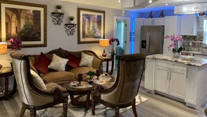 Un salon moderne avec un canapé, des fauteuils et une table basse. On y trouve également une cuisine dotée de meubles blancs et d’un réfrigérateur.