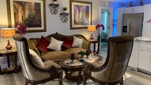 Un salon comprenant un canapé, deux fauteuils, une table basse et des objets de décoration. On y trouve deux étagères murales ornées de plantes et un tableau encadré. On aperçoit également, au fond, un coin cuisine avec un réfrigérateur.