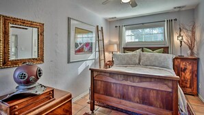 Quarto com cama de madeira, ventoinha no teto, janela com persianas, mesa de cabeceira com abajur e cômoda com rádio antigo.