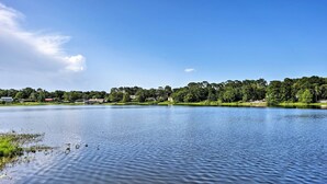 Um lago tranquilo com céu claro, cercado por vegetação e edifícios ao longe.