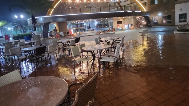 Un área de café al aire libre con mesas y sillas, un corredor cubierto y un edificio al fondo.