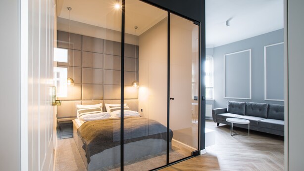 Quarto moderno com divisória de vidro, cama com coberta, mesa de cabeceira e criado-mudo.