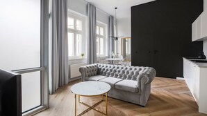 Uma sala de estar moderna com um sofá cinza com detalhes embutidos, uma mesa de centro branca redonda com pés dourados e uma janela grande com cortinas cinzas.