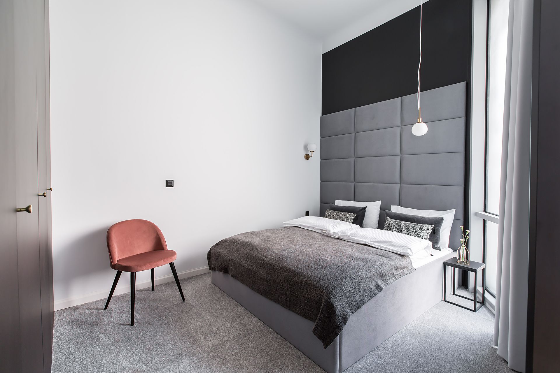 A modern bedroom with a grey upholstered headboard, a single bed with a grey blanket, a red chair, and a bedside table with a lamp.