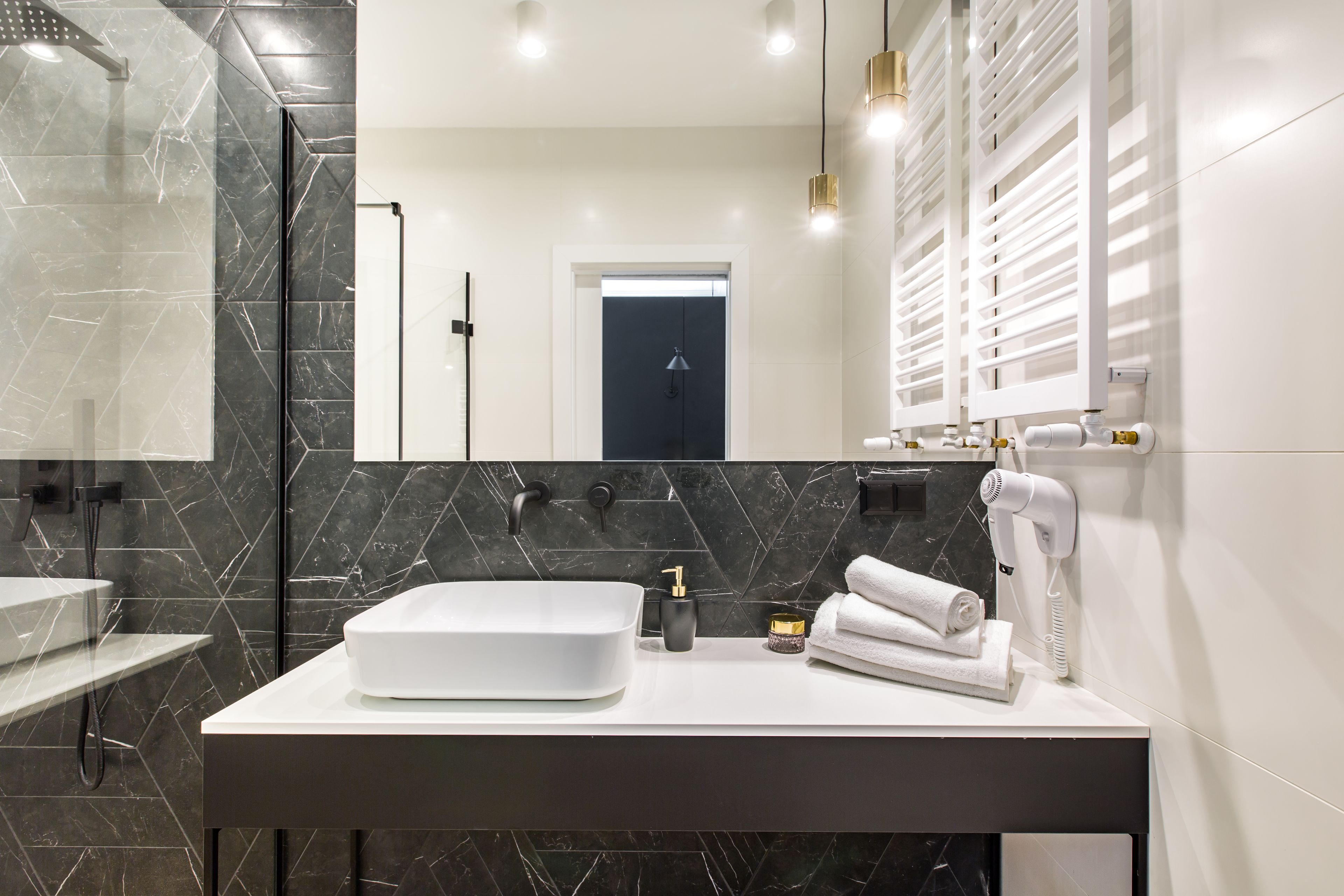 A modern bathroom with a large white sink, black marble tiles, a handheld showerhead, and a hairdryer on the counter.