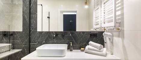 Un baño moderno con un lavabo blanco grande, azulejos de mármol negro y un cabezal de ducha manual, además de un secador de pelo en la encimera.