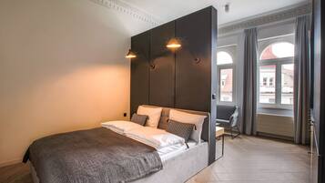 Un dormitorio moderno con una cama grande, un cabecero oscuro y vistas de la ciudad a través de amplias ventanas.