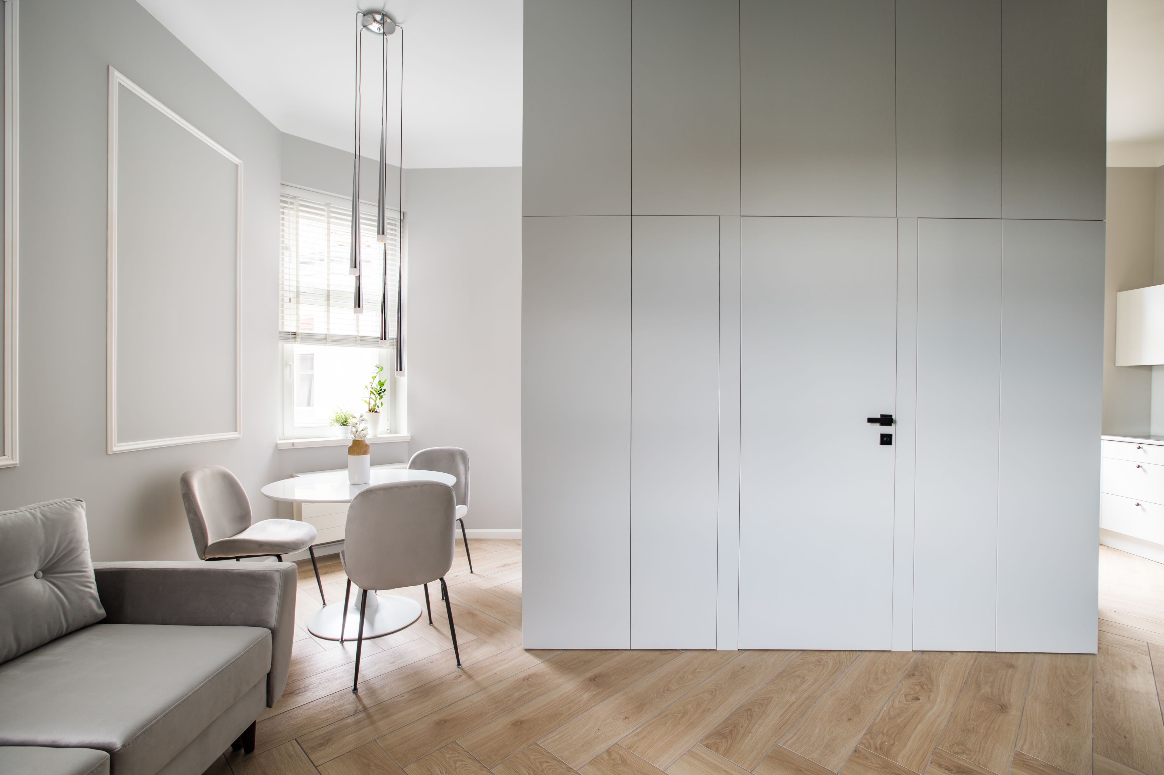 A modern living room with a grey sofa, a dining table with chairs, and a large white wardrobe.