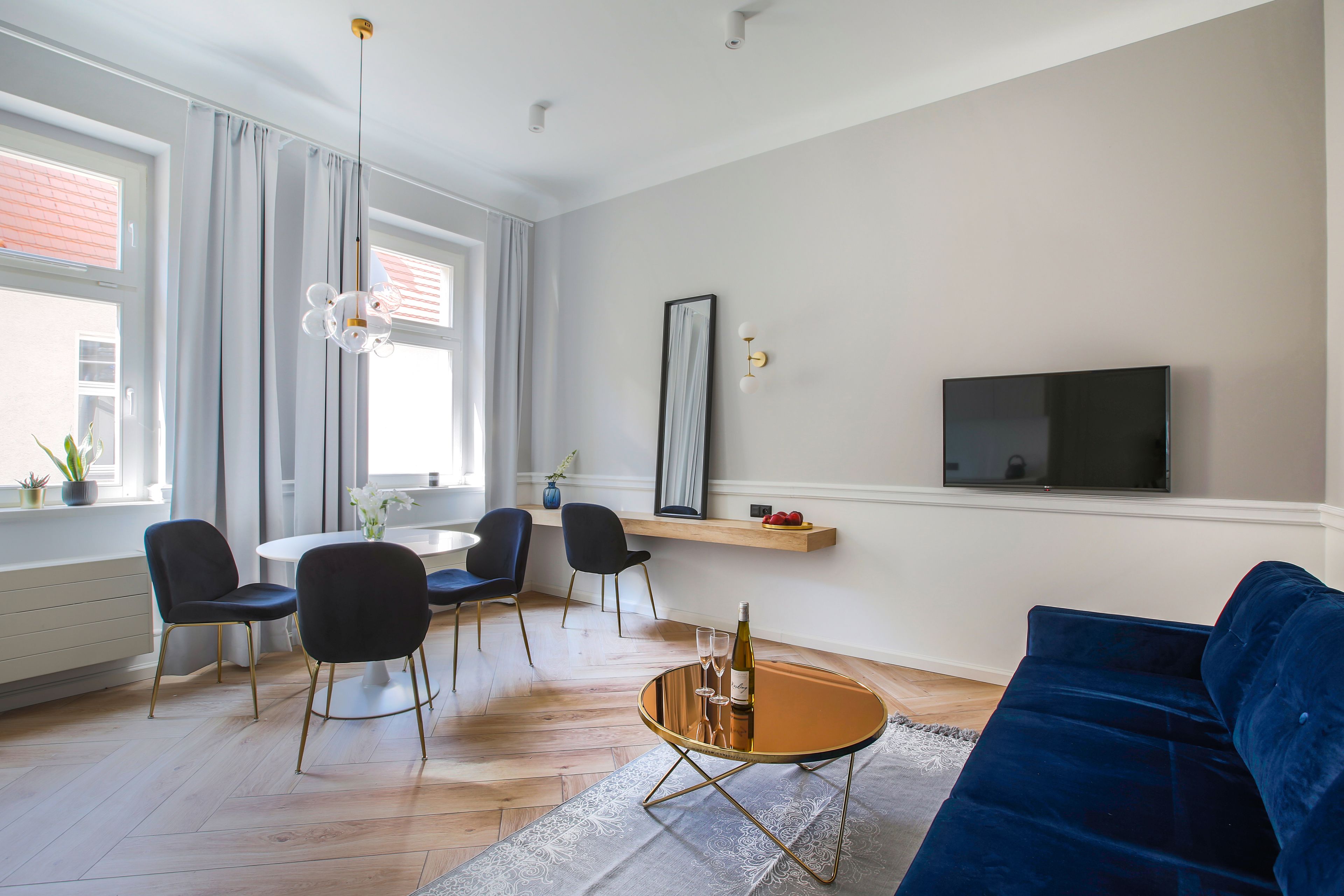 A modern living room with a blue sofa, a round coffee table with a bottle and glasses, a TV on the wall, and a dining area with chairs and a table.