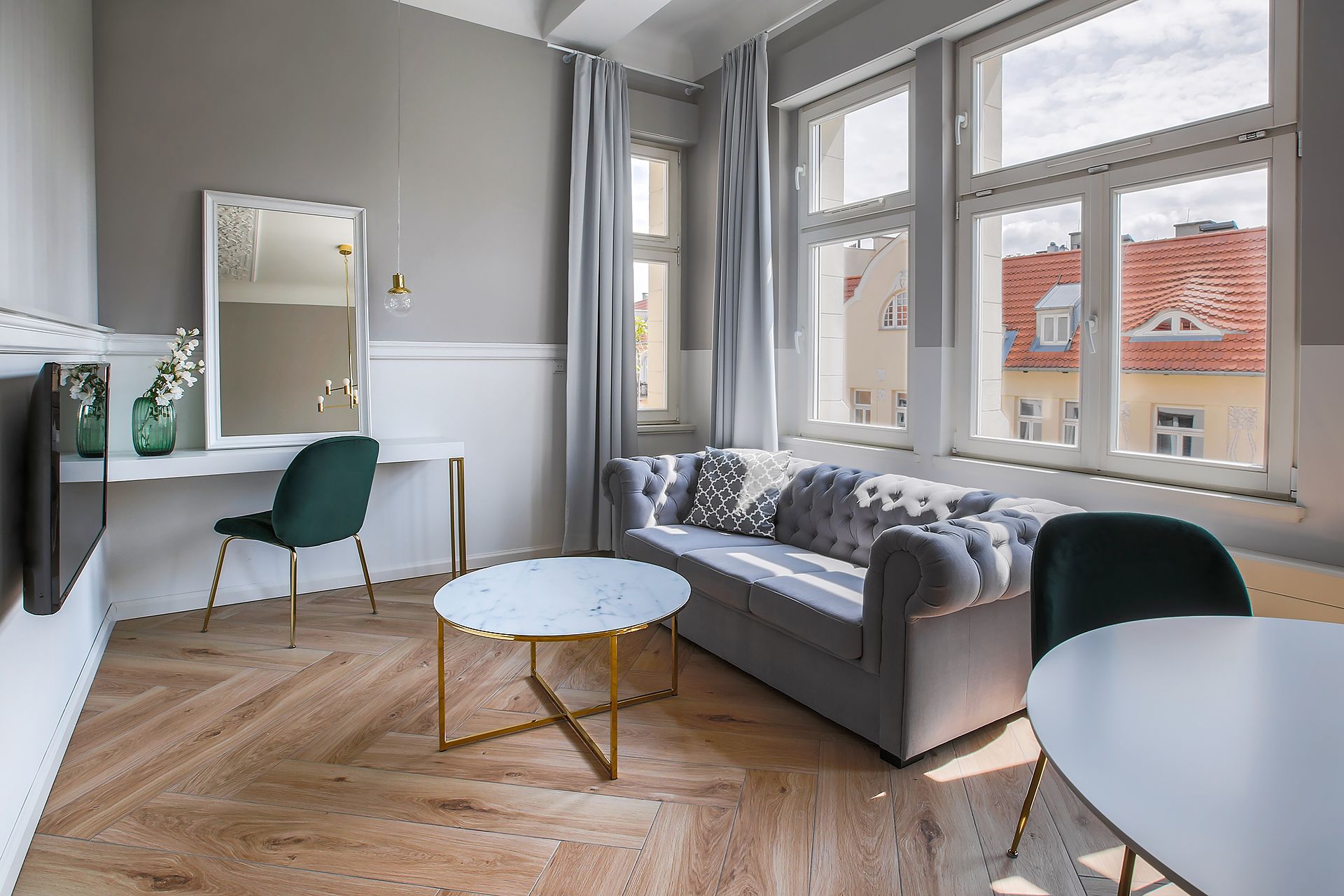 A modern living room with a grey sofa, a round coffee table, and a green chair. There is a large window with curtains, a flat-screen TV, and a mirror on the wall.