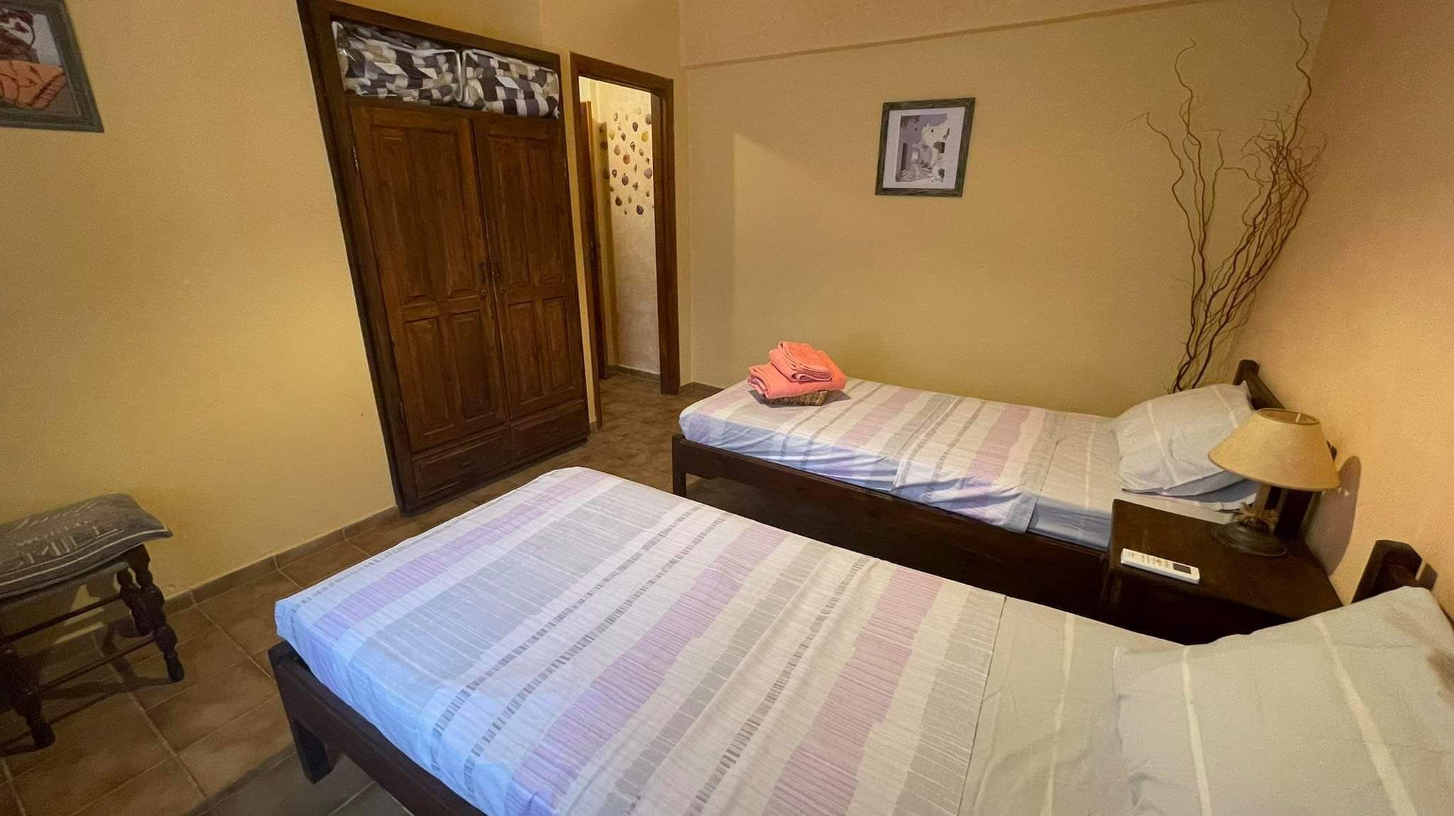 A small, simple room with two beds, a wooden wardrobe, a small table, and a lamp.