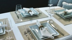 Tavola imbandita per quattro, con piatti quadrati, bicchieri da vino e posate, su un tavolo bianco con vista sul mare.