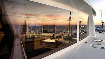 A modern yacht interior with a large glass window offering a view of the ocean and sunset.