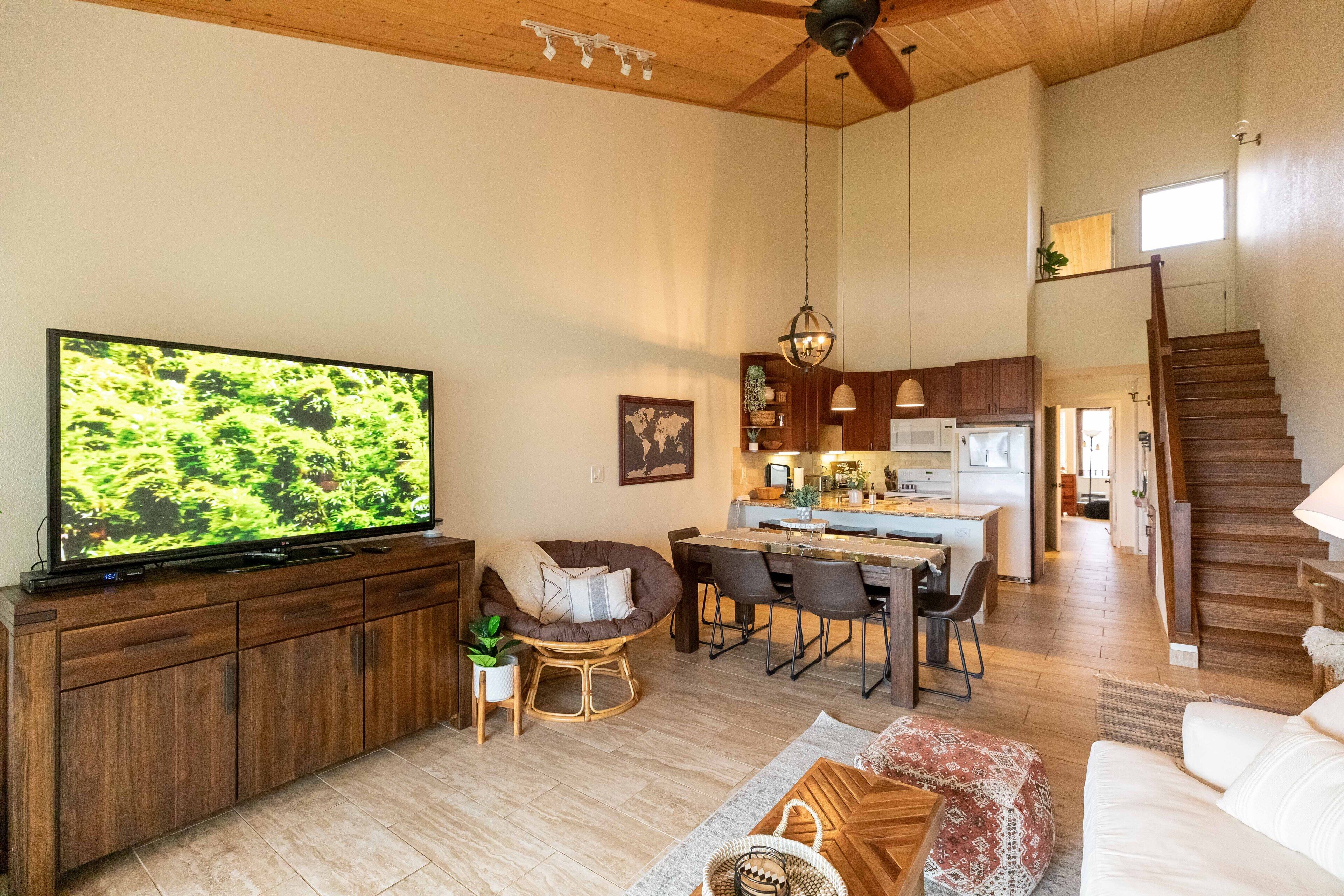 A spacious living room with a large flat-screen TV, wooden flooring, a dining area with a kitchen in the background, and a staircase leading to an upper level.