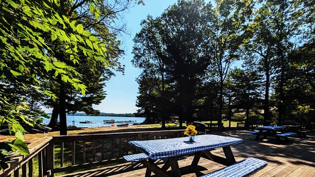 A wooden deck with picnic tables and benches, overlooking a lake with boats and a dock.