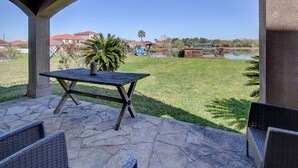 Un patio con una mesa de piedra y sillas de mimbre, con vistas a una zona verde con un lago y casas al fondo.