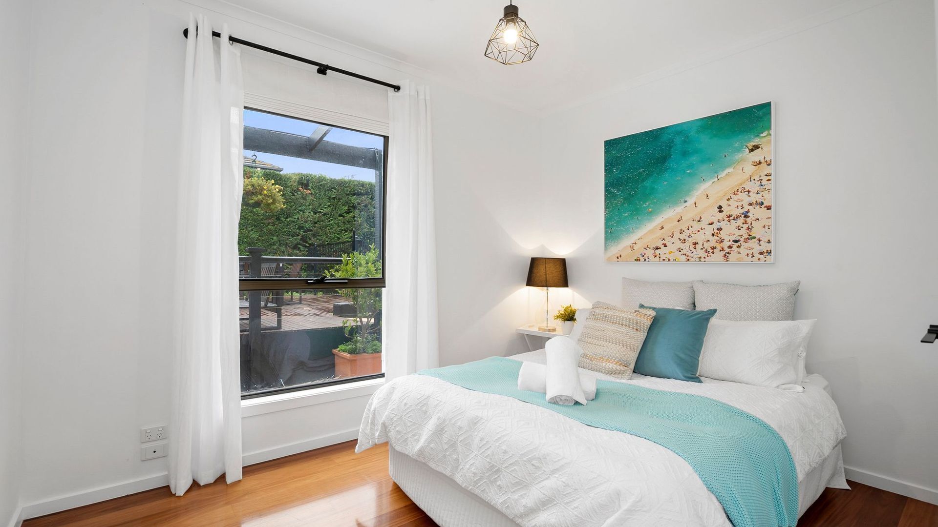 A bedroom with a large bed, a view of a balcony, a beach painting, and a pendant light.