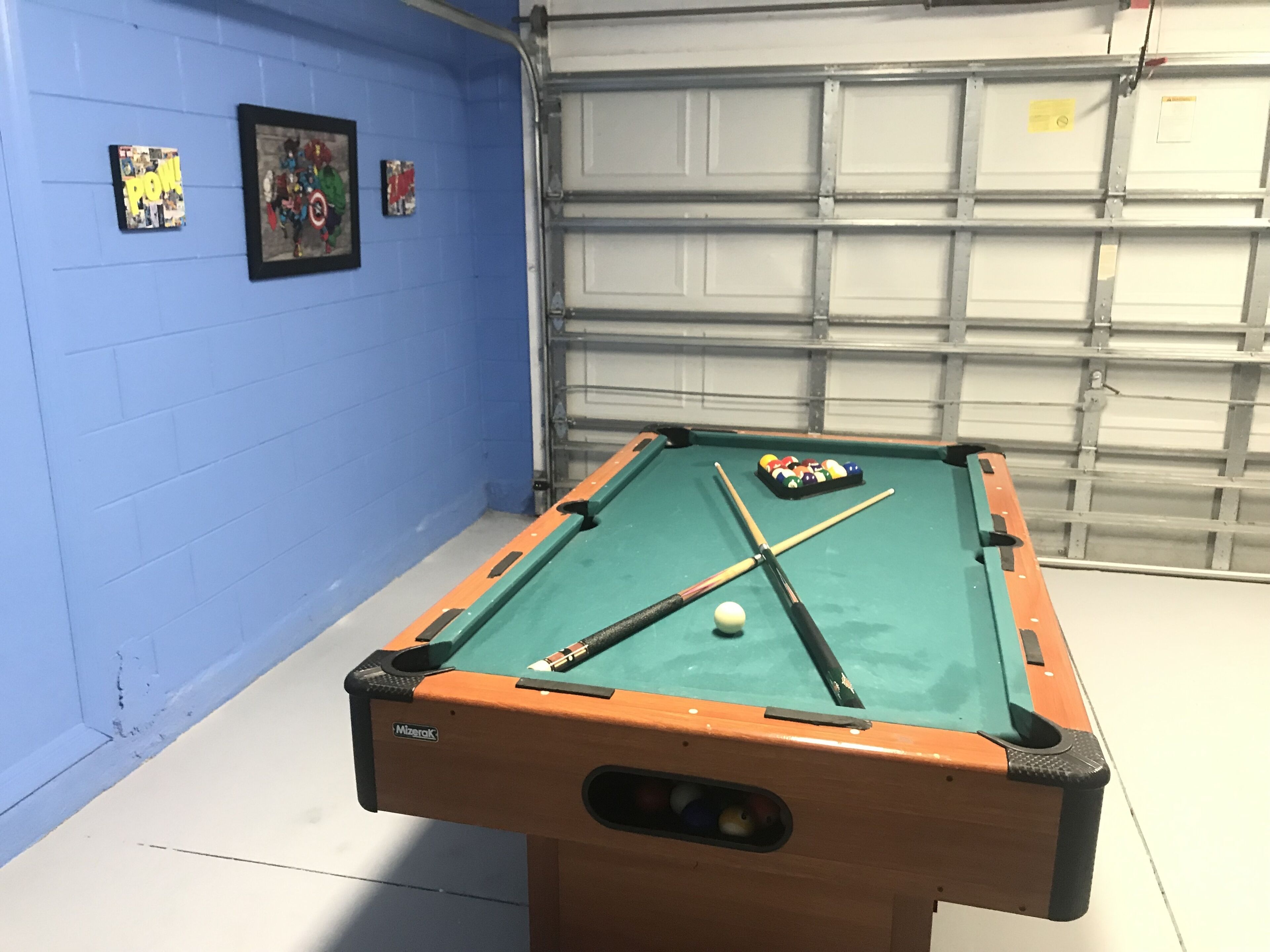 A pool table with a cue stick and balls, a blue wall with framed pictures, and a garage door in the background.